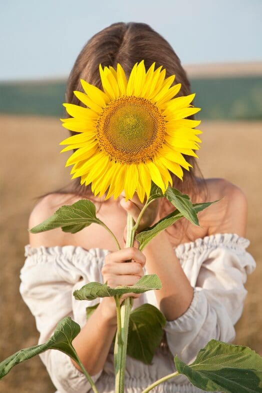 woman in a white top standing in a field holding a sunflower in front of her face